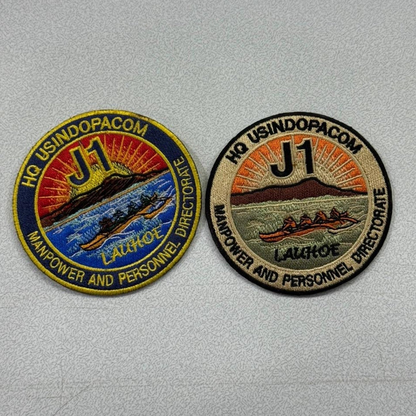 Two round embroidered patches of the same HQ USINDOPACOM J1 design side by side, one in full colour with a blue and gold border and one in a muted olive colourway, each showing a sunrise over an outrigger canoe with the words MANPOWER AND PERSONNEL DIRECTORATE and LAUHOE
