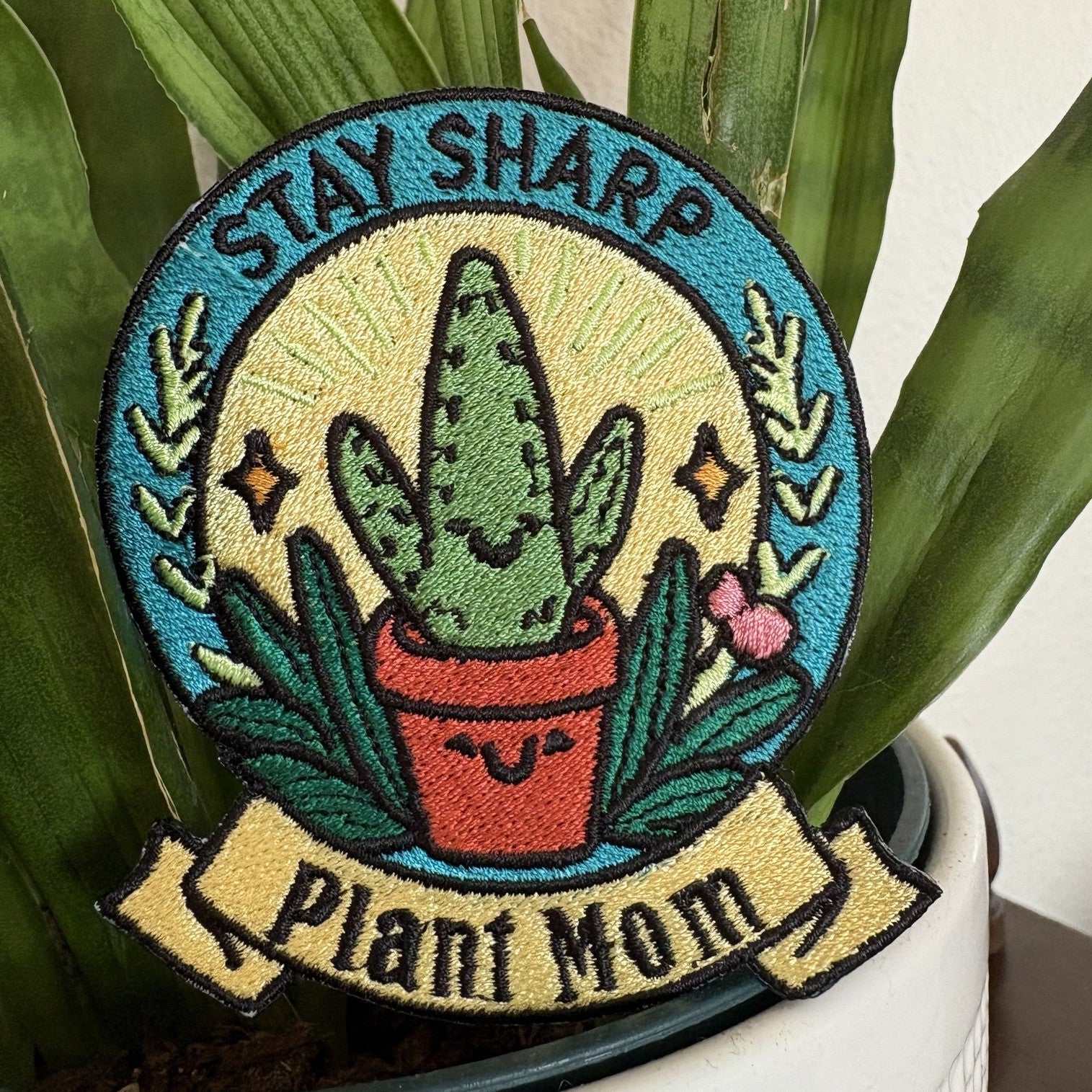 Round embroidered patch with a teal border reading STAY SHARP around a smiling potted cactus on a cream ground, above a yellow scroll banner reading Plant Mom, photographed beside a houseplant