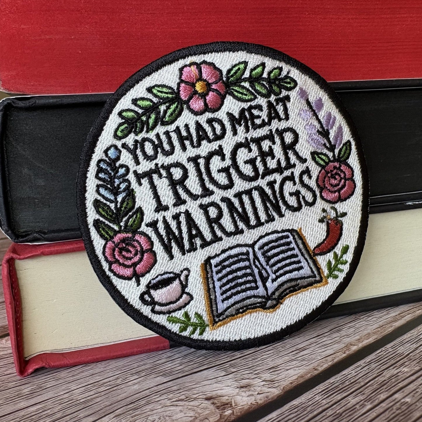 Round embroidered patch with a black border and white ground reading YOU HAD ME AT TRIGGER WARNINGS, surrounded by flowers, an open book, a teacup and a chilli, resting on a stack of books