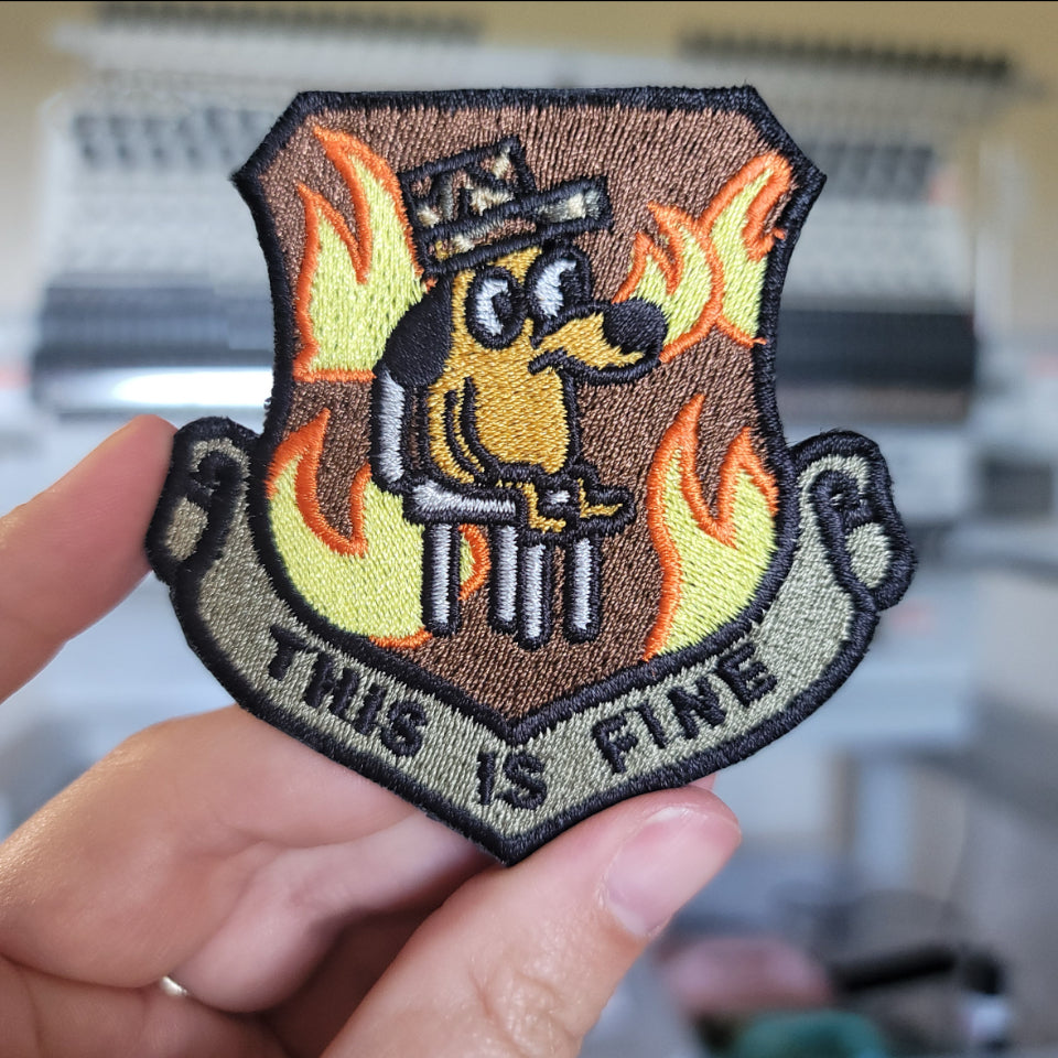 Shield-shaped embroidered morale patch held in hand in front of an embroidery machine: a cartoon dog in a peaked cap sitting calmly among yellow and orange flames, over a scroll banner reading THIS IS FINE