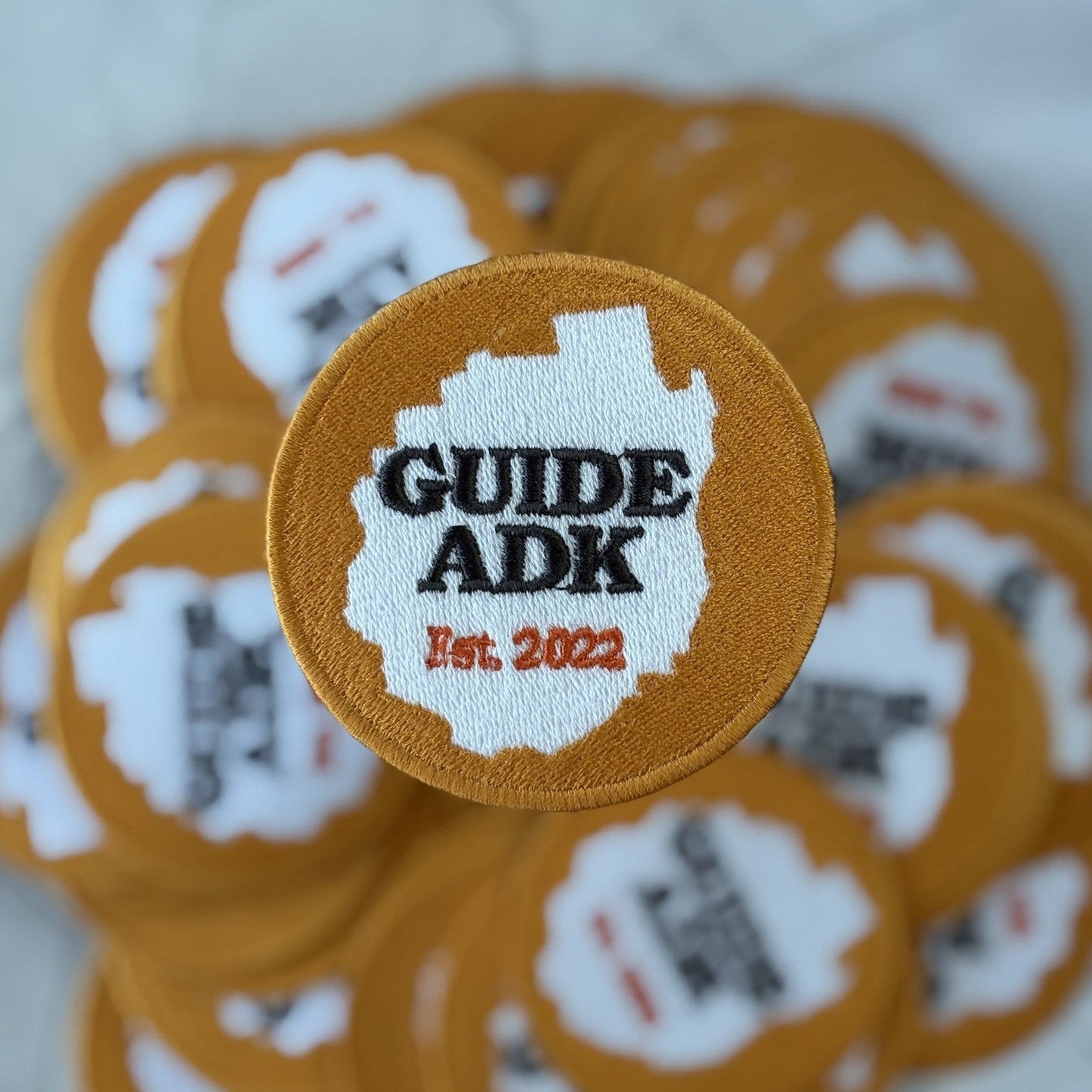 Round embroidered patch in ochre with a white silhouette of the Adirondack Park and the lettering GUIDE ADK, Est. 2022, sitting on a pile of identical patches