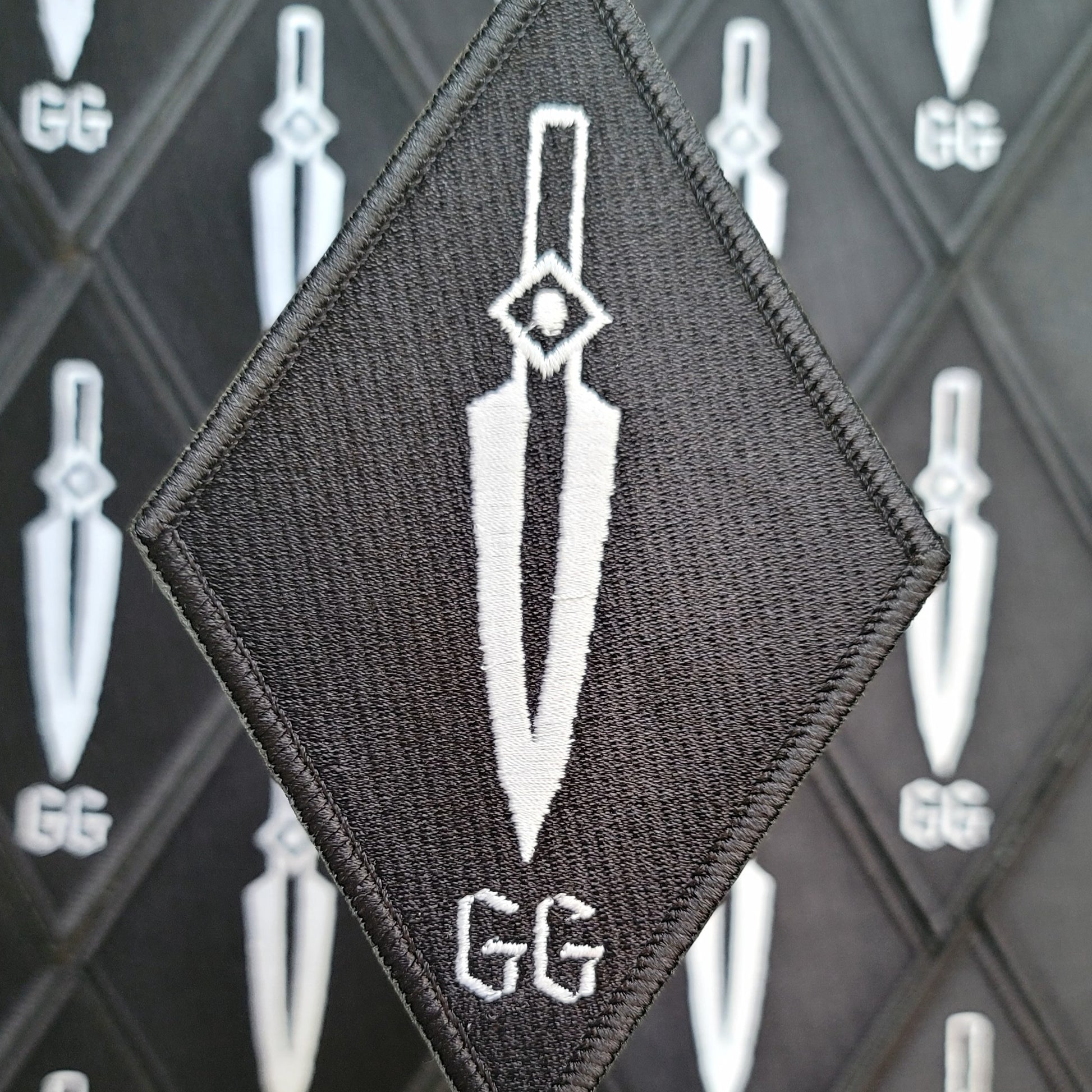 Diamond-shaped charcoal embroidered patch with a white dagger blade point-down and the letters GG beneath it, shown against a rack of identical patches