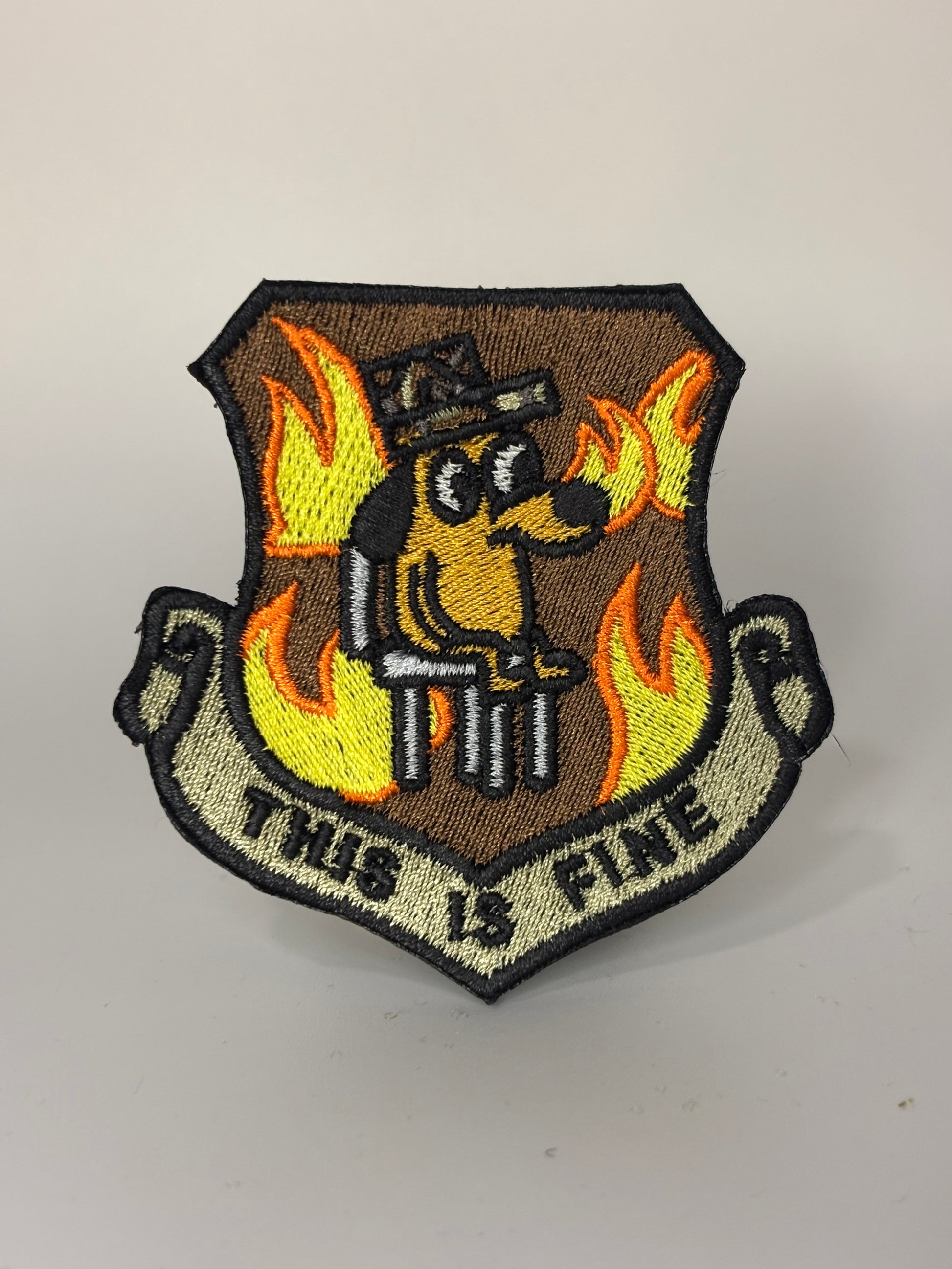 Shield-shaped embroidered morale patch in brown and olive drab with yellow and orange flames, a cartoon dog in a peaked cap, and a scroll banner reading THIS IS FINE, with a black merrowed edge